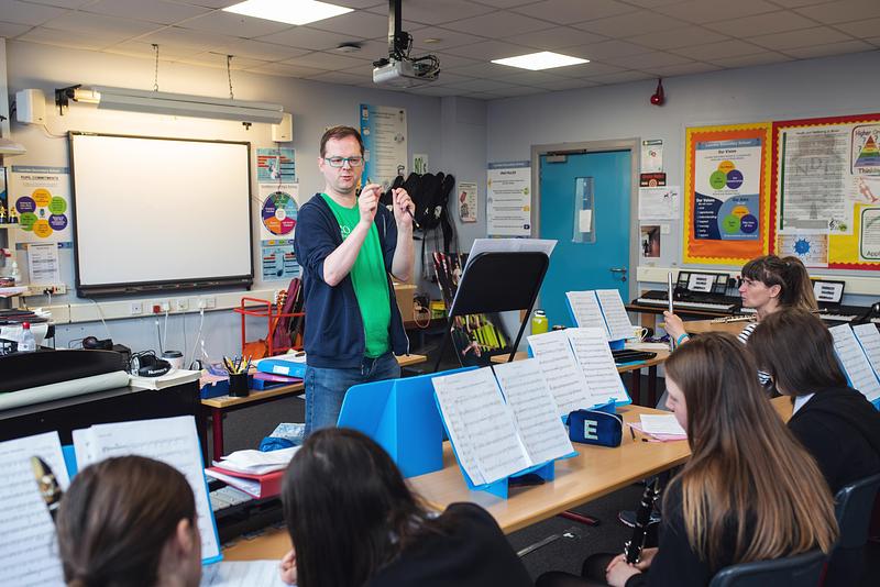 A man in a green Royal Scottish National Orchestra (RSNO) t-shirt standing in front of a classroom orchestra wind instrument section